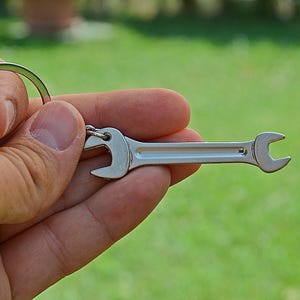 Op de afbeelding: Een zilverkleurige metalen sleutelhanger met een miniatuur steeksleutelbedel bevestigd aan een ronde splitring. De sleutel heeft een gladde, gepolijste afwerking met een centrale verzonken groef die over de lengte van de steel loopt.