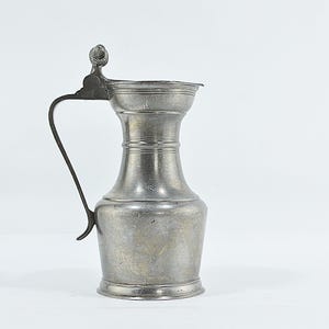 May include: A metallic grey pewter pitcher featuring a wide base, a narrow neck, and a flared rim. The vessel includes a curved handle on the side and a hinged lid with a small decorative finial at the top. The surface has a matte, aged finish.