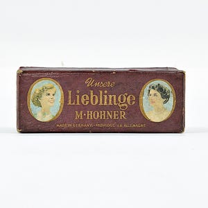 May include: A vintage maroon box with the text "Unsere Lieblinge M. Hohner" in gold lettering. The box features oval portraits of women on each end. The text "Made in Germany - Fabriqué en Allemagne" is also visible.