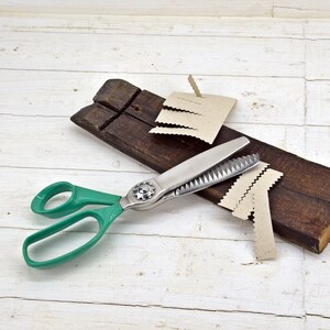 May include: Green handled pinking shears with a silver blade cutting brown paper strips on a white wooden surface.