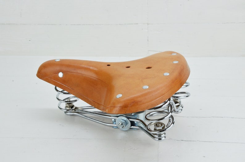 May include: A brown leather bicycle saddle with a curved design. The saddle is supported by a chrome-colored spring suspension system. The saddle is approximately 10 inches long and 8 inches wide.