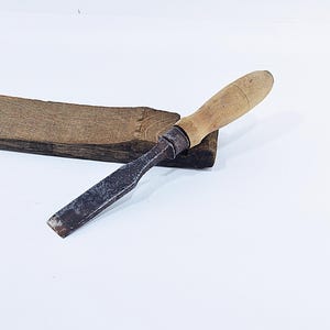 May include: A vintage wood chisel with a wooden handle and a metal blade, resting on a piece of dark brown wood. The chisel is a carpentry tool, used for shaping wood.
