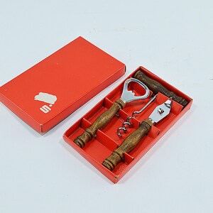 Op de afbeelding: Een vintage bar-gereedschapsset in een rode doos. De set bevat een flesopener, kurkentrekker en een blikopener, elk met houten handvatten en zilverkleurige metalen componenten. De doos heeft een wit logo op het deksel.