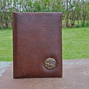 May include: A brown leather-bound book with a textured cover. A circular brass emblem with a horse and rider is affixed to the lower right corner. The book is standing upright on a weathered surface, with a green grassy background.