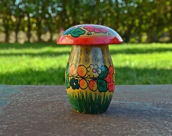 May include: A hand-painted wooden mushroom-shaped container with a yellow base, red and green accents, and floral designs. The container is sitting on a brown surface.