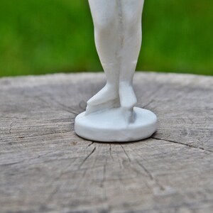 以下が含まれることがあります： 木製の表面に立っている白い陶器の人形。人形はシンプルなデザインで、繊細な線で装飾されています。