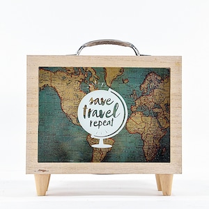May include: A wooden piggy bank shaped like a suitcase with a world map design and the text "save travel repeat" on a white circle. The piggy bank has a metal handle and wooden legs.