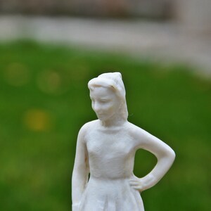 以下が含まれることがあります： 髪を後ろでまとめた女の子の白い磁器の置物。シンプルなドレスを着て、物思いにふけっている表情をしています。