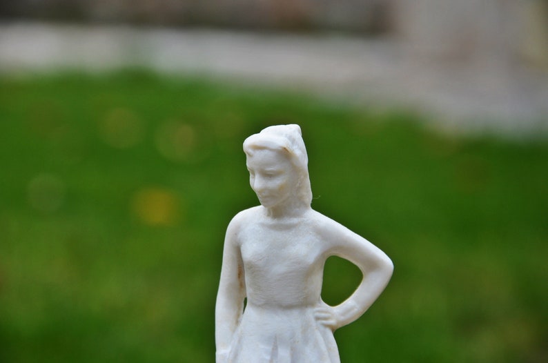 以下が含まれることがあります： 髪を後ろでまとめた女の子の白い磁器の置物。シンプルなドレスを着て、物思いにふけっている表情をしています。