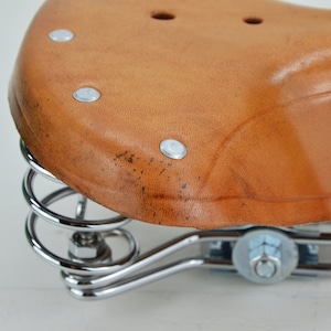 May include: Close-up of a brown leather bicycle seat with a curved design. The seat is attached to a chrome-colored spring suspension system. The leather has a slightly worn appearance.