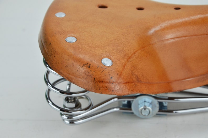 May include: Close-up of a brown leather bicycle seat with a curved design. The seat is attached to a chrome-colored spring suspension system. The leather has a slightly worn appearance.