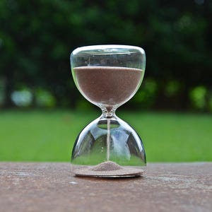 May include: A clear glass hourglass with brown sand inside. The hourglass is set against a blurred green background of trees and grass. The sand is flowing from the top to the bottom chamber, indicating the passage of time.