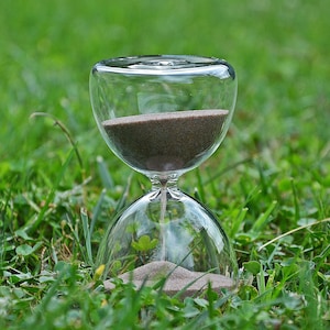 Op de afbeelding: Een glazen zandloper met bruin zand op een bedje van groen gras.