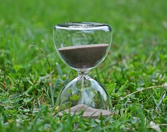 May include: A glass hourglass with brown sand sitting on a bed of green grass.