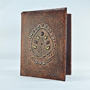 May include: A rectangular brown journal with a textured, pebbled surface. The front cover features an embossed, teardrop-shaped design decorated with small, inlaid yellow and dark red stones, surrounded by a border of tiny silver-toned metal studs.