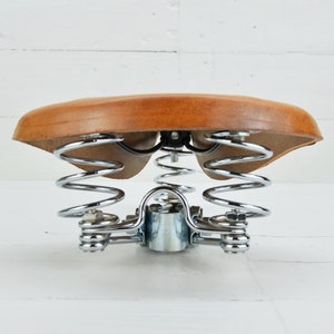 May include: A vintage bicycle saddle with a light brown leather seat and chrome-plated springs. The seat is suspended by two sets of springs, providing a comfortable ride. The saddle is designed for bicycles.