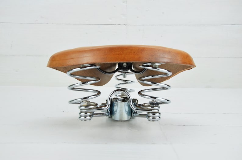 May include: A vintage bicycle saddle with a light brown leather seat and chrome-plated springs. The seat is suspended by two sets of springs, providing a comfortable ride. The saddle is designed for bicycles.