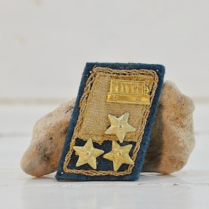 May include: A vintage military insignia, featuring a rectangular navy blue base with gold trim. The insignia has a gold emblem at the top and three gold stars. The item is displayed on a light brown stone.
