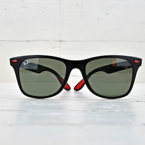 K&ouml;nnte beinhalten: Schwarze Sonnenbrille mit roten Akzenten an den B&uuml;geln und gr&uuml;nen Gl&auml;sern. Die Sonnenbrille liegt auf einer wei&szlig;en Holzoberfl&auml;che.