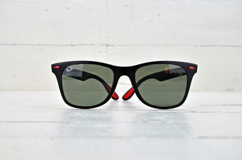 K&ouml;nnte beinhalten: Schwarze Sonnenbrille mit roten Akzenten an den B&uuml;geln und gr&uuml;nen Gl&auml;sern. Die Sonnenbrille liegt auf einer wei&szlig;en Holzoberfl&auml;che.