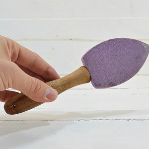 Puede incluir: Un cepillo de mango de madera con una cabeza de esponja morada. El cepillo se sostiene en una mano.