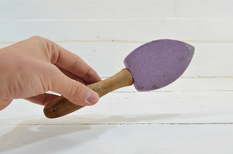 Puede incluir: Un cepillo de mango de madera con una cabeza de esponja morada. El cepillo se sostiene en una mano.