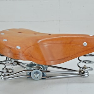 May include: A brown leather bicycle saddle with a curved shape and silver rivets. The saddle is supported by two chrome-plated springs and a chrome frame. The design is vintage and classic.
