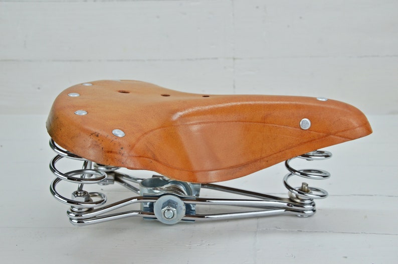 May include: A brown leather bicycle saddle with a curved shape and silver rivets. The saddle is supported by two chrome-plated springs and a chrome frame. The design is vintage and classic.