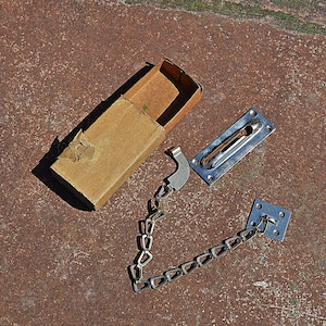 May include: A vintage door chain lock set with a cardboard box. The set includes a metal chain, two metal plates, and a latch. The metal components have a silver finish. The box is brown.