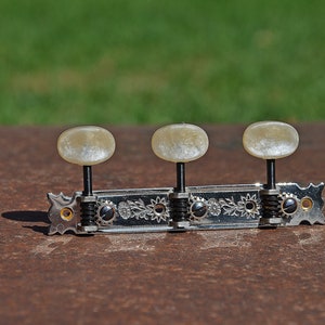 Puede incluir: Primer plano de tres clavijas de afinación plateadas con pomos de plástico blanco en una guitarra. Las clavijas están unidas a una placa de metal con diseños florales decorativos.