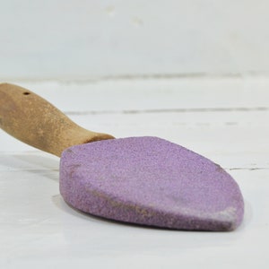 Puede incluir: Una piedra de afilar de color morado con un mango de madera. La piedra tiene forma de pala peque&ntilde;a.