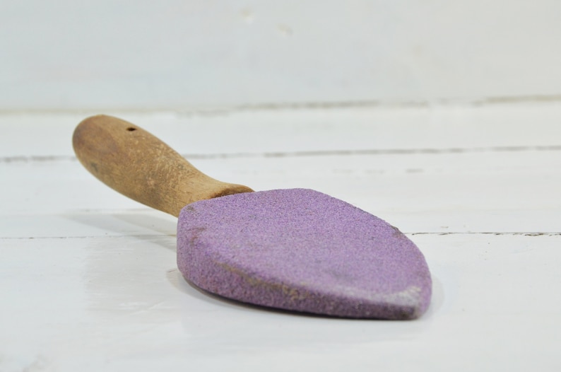 Puede incluir: Una piedra de afilar de color morado con un mango de madera. La piedra tiene forma de pala peque&ntilde;a.