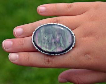 Puede incluir: Un anillo de plata con una gran piedra ovalada iridiscente engastada en el centro. La piedra tiene un patrón de remolino de verde, morado y negro.