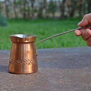 Può includere: Una caffettiera di rame con un manico lungo. La caffettiera ha un design intricato e si trova su una superficie marrone.