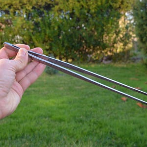 May include: A pair of stainless steel tweezers, held against a backdrop of green grass and foliage. The tweezers are long, with textured grips for precision. The tips are pointed, designed for fine manipulation.