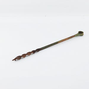May include: A vintage auger drill bit with a spiral cutting edge and a loop handle. The metal tool has a rusty brown and green patina, suggesting age and use. The auger is designed for boring holes in wood.