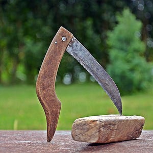 Może przedstawiać: Składana piła z zakrzywionym, zniszczonym metalowym ostrzem i teksturowaną brązową drewnianą rękojeścią. Piła jest ustawiona tak, że jej końcówka opiera się na prostokątnym, jasnobrązowym drewnianym klocku, co nadaje jej rustykalny i zużyty wygląd.