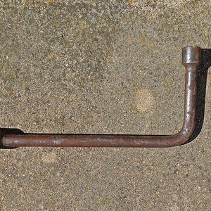 Puede incluir: Una llave de tuercas en forma de L oxidada. La herramienta está hecha de metal y tiene un casquillo en cada extremo. La llave está sobre una superficie de hormigón rugosa y texturizada.