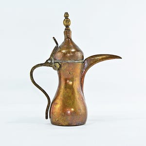 May include: A metallic coffee pot with a warm bronze and copper patina, featuring a tall, slender body, a curved handle, a long pointed spout, and a removable lid topped with a small, decorative finial. The surface shows signs of age.