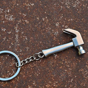 Op de afbeelding: Een zilverkleurige metalen sleutelhanger met een miniatuur hamerbedel die met een korte metalen ketting aan een ronde splitring is bevestigd. De hamerbedel heeft een gladde, gepolijste afwerking en een klassiek ontwerp met een hamerkop.