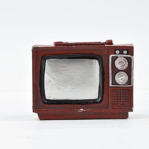 Puede incluir: Un televisor de plástico marrón de estilo vintage con una pantalla plateada. El televisor tiene dos botones redondos en el lado derecho y un patrón texturizado en la parte inferior.