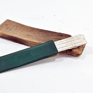 Puede incluir: Una regla de cálculo de plástico blanco con marcas numéricas y escalas negras, parcialmente insertada en una funda de cuero texturizado de color verde oscuro. La regla presenta el texto "ARISTO" y "Nr. 0968" impreso en tinta negra.