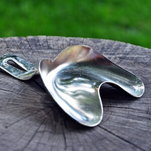 Può includere: Una paletta in metallo argentato lucido, con un design curvo e un manico con un foro. È appoggiata su una superficie di legno grigio e usurato, con uno sfondo verde sfocato.