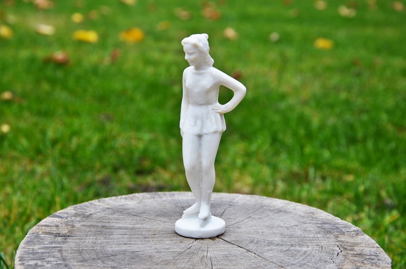 以下が含まれることがあります： 片手に腰を当てて立っている女性の白い磁器製フィギュア。フィギュアは木製の台座の上に置かれています。