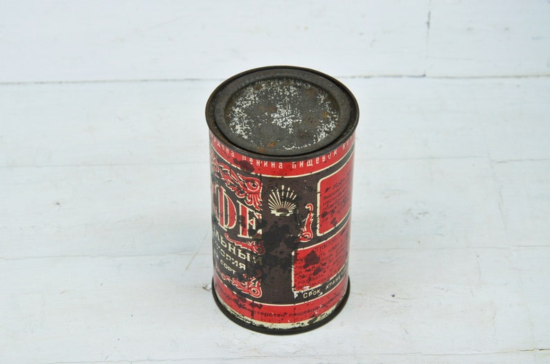 May include: A vintage metal can with a red and black label. The label features the text "Кофе" in Cyrillic script, which translates to "Coffee". The can is slightly rusted and shows signs of wear.