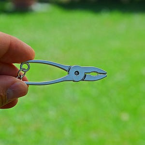 May include: A miniature silver-coloured metal pliers keyring featuring a small circular ring attached to the handle. The tool has a smooth, polished finish with a serrated jaw design and a central pivot point, presented as a functional accessory.