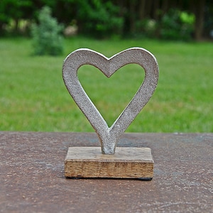 Op de afbeelding: Een rustieke metalen hartsculptuur met een getextureerde, zilvergrijze afwerking. De hartvorm is open in het midden en gemonteerd op een kleine, rechthoekige houten basis. De sculptuur wordt buiten tentoongesteld tegen een achtergrond van groen gras en bomen.