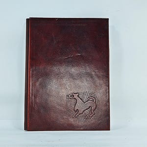 Puede incluir: Un diario rectangular de cuero marrón oscuro con un acabado liso y pulido. La portada presenta un gráfico grabado de un animal estilizado de cuatro patas sobre un patrón geométrico similar a una red en la esquina inferior derecha.