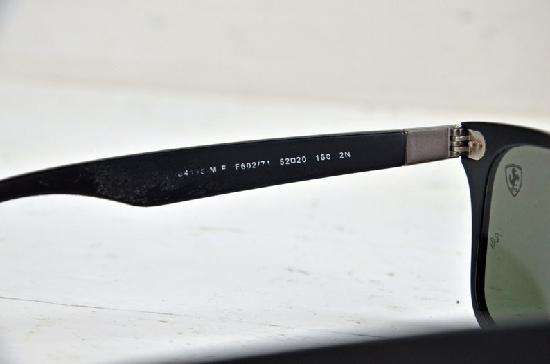 K&ouml;nnte beinhalten: Schwarze Sonnenbrille mit gr&uuml;nen Gl&auml;sern und einem silbernen Ferrari-Logo an der Seite. Die Sonnenbrille hat den folgenden Text am B&uuml;gel: "C4115 M E F602/71 52□20 150 2N"