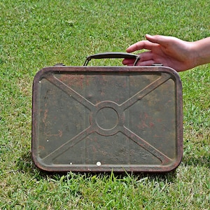 May include: A rectangular, olive green metal lunch box featuring a central circular embossed design with diagonal lines extending to the corners. The surface shows signs of rust and wear, and it includes a dark metal handle on the top edge.